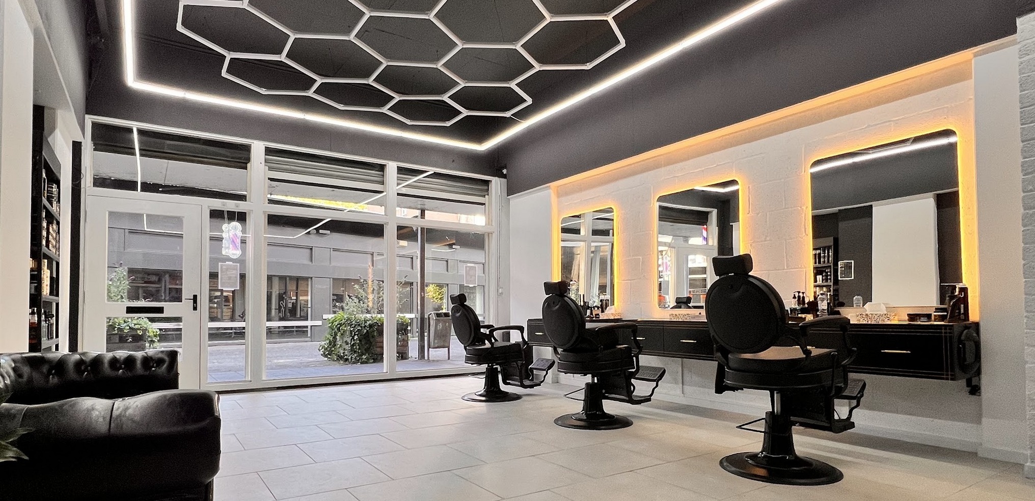 Het interieur van Nova Barbershop: honingraatverlichting aan het plafond, verlichte spiegels en barbierstoelen, met de glazen pui aan de voorkant