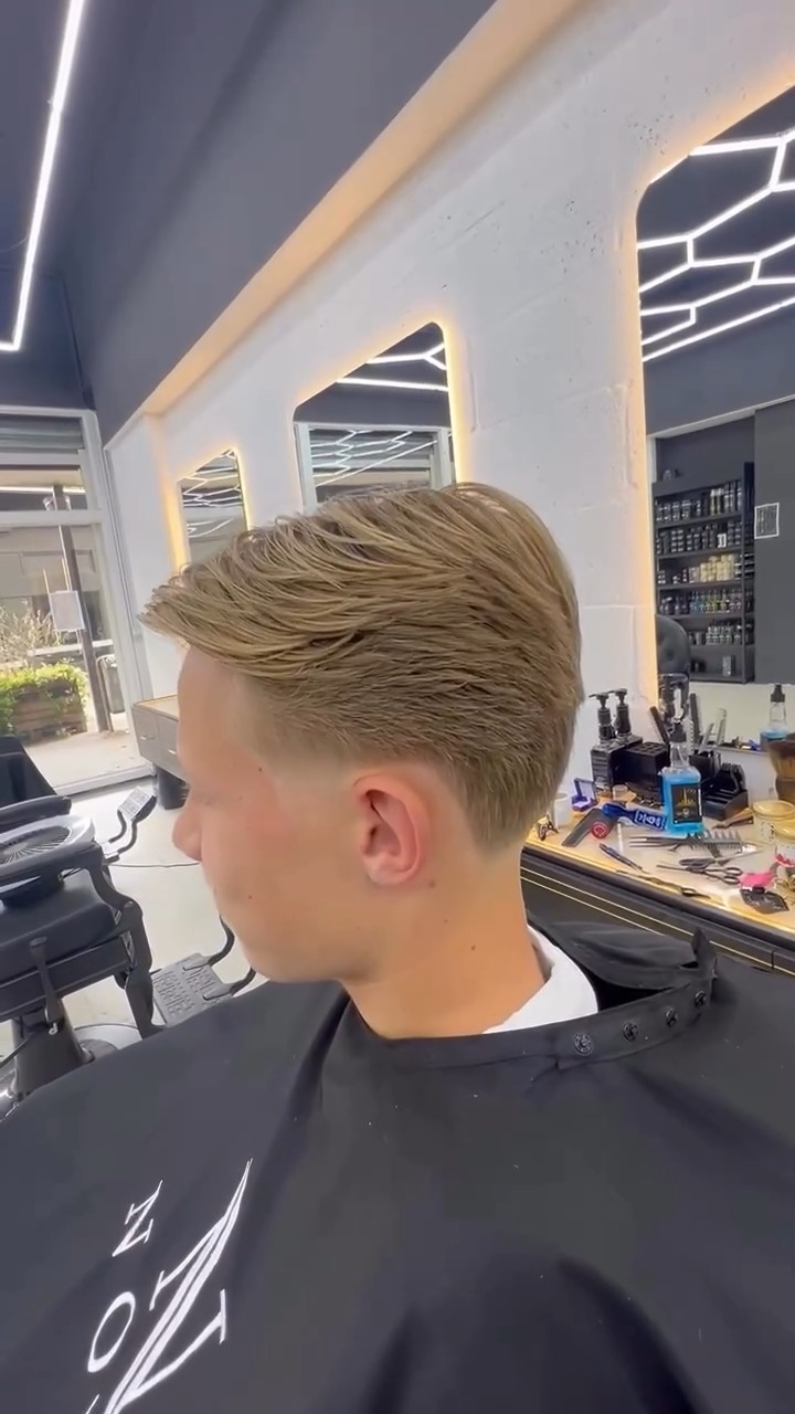 Zijaanzicht van een jonge klant met een fade en een lange blonde top, in de zaak